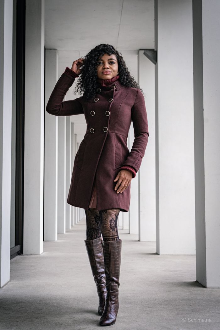 who-we-are Stylish woman in a maroon coat poses confidently in a modern urban corridor.